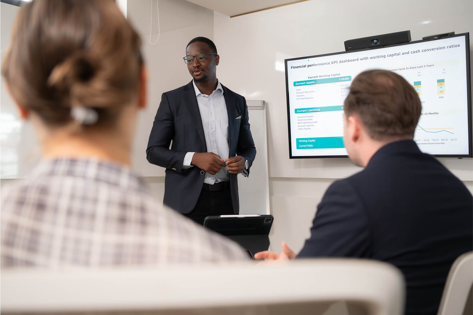 Business presenter in a suit delivering a financial performance KPI presentation to colleagues, with an interactive dashboard on working capital and cash conversion ratios displayed on a screen as part of Spotfire data visualization training.